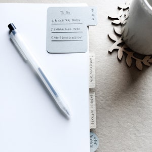 May include: A white folder with gray tabs labeled "To Do", "Jahresplan 2014", "Aktuelle Aufträge", and "Neue Produkte". The folder has handwritten notes on the tabs. The folder is on a light brown surface.