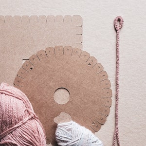 Op de afbeelding: Een close-up van een knutselset voor het maken van vriendschapsarmbanden. De set bevat twee kartonnen sjablonen, een ronde en een vierkante, met nummers die rond de randen zijn gedrukt. Er zijn ook twee bollen garen, een roze en een witte, en een roze koord.