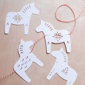 May include: A string of white paper cut-out horses, known as Dala horses, are strung together with red and white twine. The horses have intricate cut-out designs.