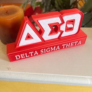 Może przedstawiać: Czerwony blok wydrukowany w 3D z literami "A&Sigma;&Theta;" w kolorze białym i słowami "Delta Sigma Theta" w kolorze białym pod literami.