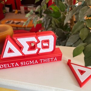 Może przedstawiać: Czerwono-białe litery wydrukowane w 3D, tworzące napis "A&Sigma;&Theta;" z napisem "Delta Sigma Theta" poniżej.