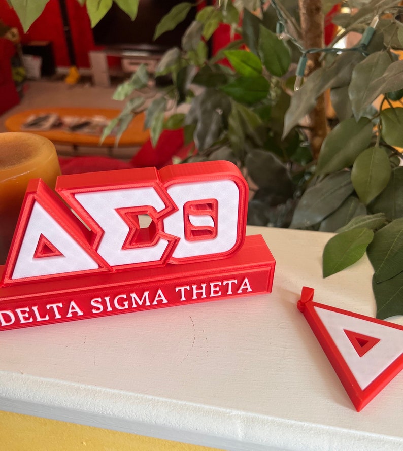 Może przedstawiać: Czerwono-białe litery wydrukowane w 3D, tworzące napis "A&Sigma;&Theta;" z napisem "Delta Sigma Theta" poniżej.