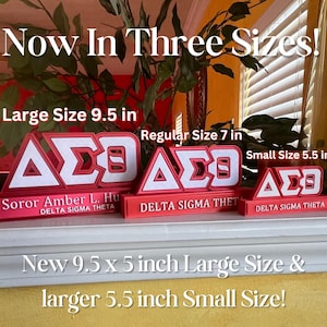 Może przedstawiać: Trzy rozmiary czerwono-białych tabliczek sororities Delta Sigma Theta z literami "&Delta;&Sigma;&Theta;" w kolorze białym. Tabliczki znajdują się na p&oacute;łce z tekstem "Now In Three Sizes!" nad nimi. Największa tabliczka ma 9,5 cala, środkowa 7 cali, a najmniejsza 5,5 cala.