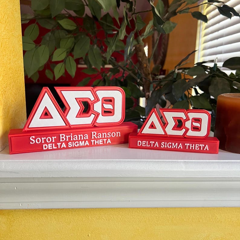 Może przedstawiać: Dwie czerwono-białe tabliczki sorority Delta Sigma Theta. Większa tabliczka ma napis "Soror Briana Ranson Delta Sigma Theta" na czerwonej podstawie. Mniejsza tabliczka ma napis "Delta Sigma Theta" na czerwonej podstawie.