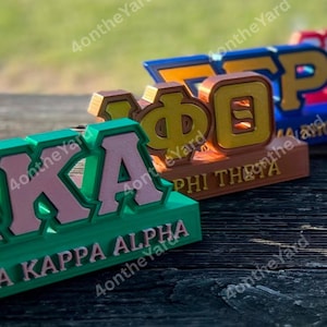 以下が含まれることがあります： ギリシャ文字の友愛会と女子学生団体の3次元サインのクローズアップ。最初のサインは緑色で、ピンク色の文字で「KAPPA ALPHA」と書かれています。2番目のサインは茶色で、黄色の文字で「PHI THETA」と書かれています。3番目のサインは青とオレンジ色です。