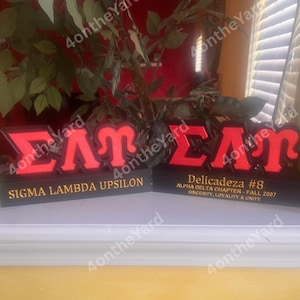 May include: Two black and red decorative signs. The first sign reads "SIGMA LAMBDA UPSILON" in gold lettering. The second sign reads "Delicadeza #8 ALPHA DELTA CHAPTER - FALL 2007 SINCERITY, LOYALTY & UNITY".