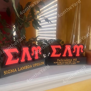 Op de afbeelding: Twee verlichte rode en zwarte Sigma Lambda Upsilon broederschapborden. Eén bord leest "Sigma Lambda Upsilon" in gouden letters. Het andere bord leest "Delicadeza #8" met "Alpha Delta Chapter - Fall 2007" en "Sincerity, Loyalty & Unity".