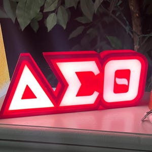 Puede incluir: Un letrero iluminado rojo y blanco que muestra las letras griegas Delta, Sigma y Theta. Las letras est&aacute;n delineadas en rojo con centros blancos iluminados, creando un contraste llamativo. El letrero es probablemente para una hermandad u organizaci&oacute;n.