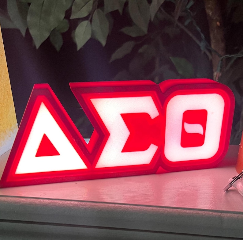 Puede incluir: Un letrero iluminado rojo y blanco que muestra las letras griegas Delta, Sigma y Theta. Las letras est&aacute;n delineadas en rojo con centros blancos iluminados, creando un contraste llamativo. El letrero es probablemente para una hermandad u organizaci&oacute;n.