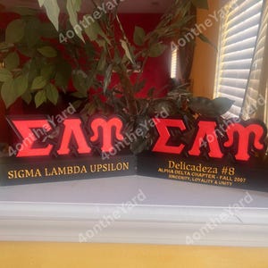May include: Two black and red Sigma Lambda Upsilon fraternity signs. The signs have the Greek letters &Sigma;&Lambda;&Upsilon; in red, with the words "Sigma Lambda Upsilon" and "Delicadeza #8" in gold lettering. The signs are displayed on a white surface.
