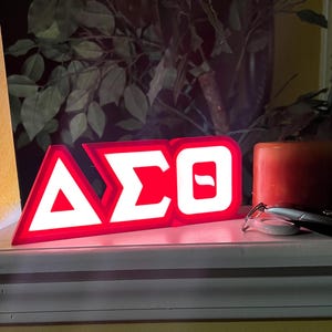 Puede incluir: Un letrero rojo y blanco iluminado que muestra las letras griegas &Delta;&Sigma;&Theta;. El letrero es rectangular con un borde blanco y las letras son blancas. Una vela roja y gafas est&aacute;n al lado del letrero.
