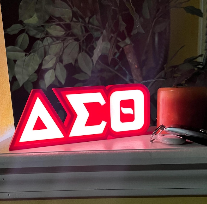 Puede incluir: Un letrero rojo y blanco iluminado que muestra las letras griegas &Delta;&Sigma;&Theta;. El letrero es rectangular con un borde blanco y las letras son blancas. Una vela roja y gafas est&aacute;n al lado del letrero.