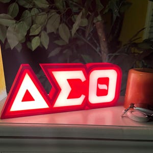 Puede incluir: Un letrero iluminado rojo y blanco que muestra las letras griegas Delta, Sigma y Theta. El letrero est&aacute; sobre una superficie blanca, con una peque&ntilde;a vela naranja y llaves en el fondo. Es probable que el letrero sea un art&iacute;culo decorativo.