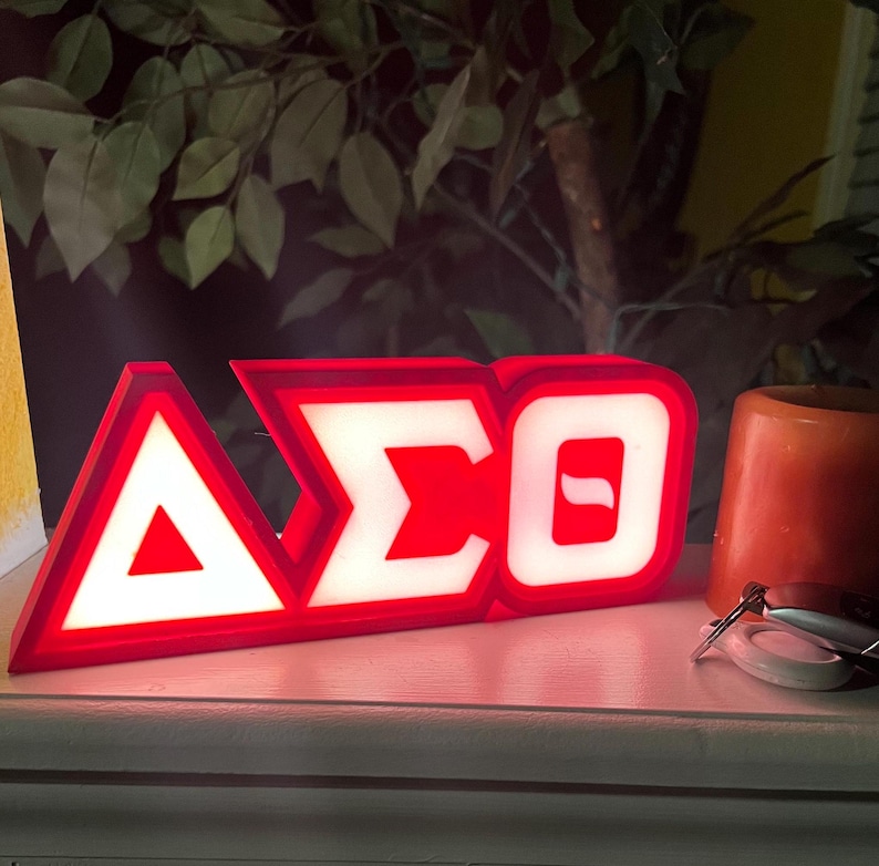 Puede incluir: Un letrero iluminado rojo y blanco que muestra las letras griegas Delta, Sigma y Theta. El letrero est&aacute; sobre una superficie blanca, con una peque&ntilde;a vela naranja y llaves en el fondo. Es probable que el letrero sea un art&iacute;culo decorativo.