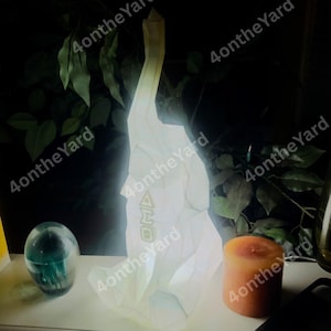 May include: A glowing white geometric sculpture with the letters "AMO" on its side. A teal glass sphere and an orange candle are next to the sculpture. The background features green foliage.