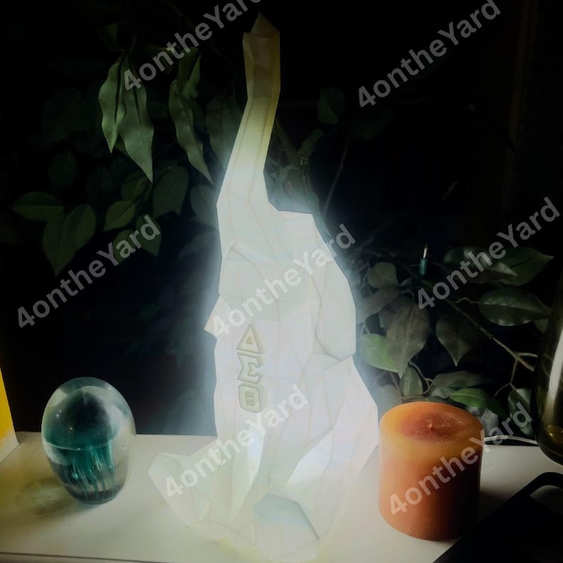 May include: A glowing white geometric sculpture with the letters "AMO" on its side. A teal glass sphere and an orange candle are next to the sculpture. The background features green foliage.
