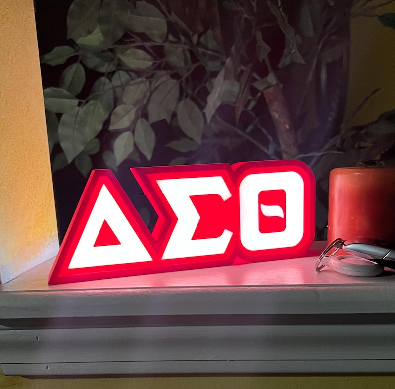 Puede incluir: Un letrero iluminado rojo y blanco que muestra las letras griegas Delta, Sigma y Theta. El letrero est&aacute; sobre una superficie blanca, con una vela roja y gafas en el fondo. El letrero es un art&iacute;culo decorativo.