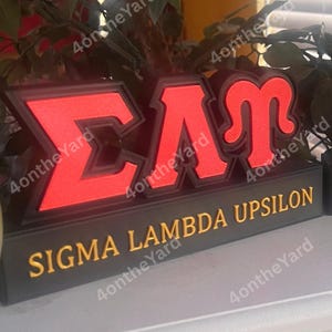 May include: A red and black Sigma Lambda Upsilon sign. The letters are in a bold, blocky font, with a black outline. The words "SIGMA LAMBDA UPSILON" are in gold lettering on a black base.