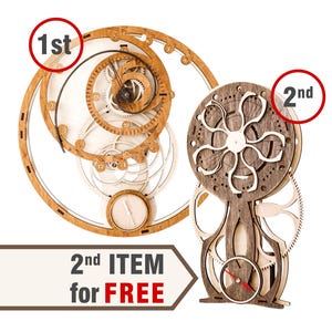May include: Two wooden mechanical models, one circular with gears and a spiral design, the other a vertical structure with a floral-like top and a clock face. The text reads "2nd ITEM for FREE".
