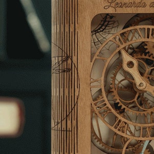 May include: A wooden clock with intricate gears and a face that reads "Leonardo da Vinci". The clock is made of light brown wood and has a detailed design.