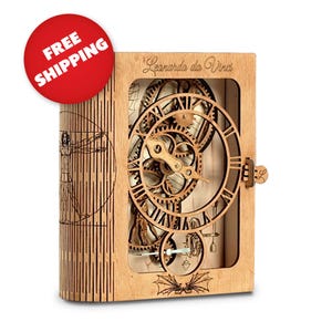 Op de afbeelding: Een houten, boekvormige klok met een wijzerplaat en ingewikkelde tandwielen zichtbaar door een venster. De klok is gemaakt van lichtbruin hout en heeft de tekst "Leonardo da Vinci" gegraveerd op de voorkant.