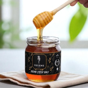 May include: A glass jar filled with golden honey, with a wooden honey dipper. The jar has a black label with white text and a floral design. Honey drips from the dipper. The jar sits on a beige cloth.