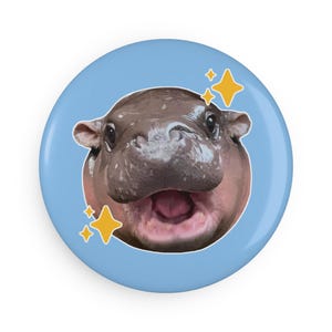 May include: A close-up of a baby hippopotamus with a wide open mouth, showing its teeth. The hippo is surrounded by a light blue background with yellow stars.