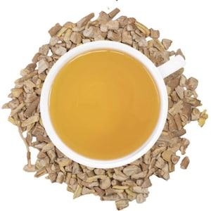 Puede incluir: Una taza blanca llena de t&eacute; dorado, rodeada de una cama de ra&iacute;z de jengibre seca y picada. El t&eacute; tiene un aspecto suave y acogedor, y los trozos de jengibre son una mezcla de marr&oacute;n claro y oscuro.