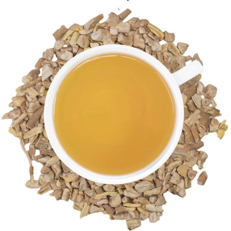 Puede incluir: Una taza blanca llena de t&eacute; dorado, rodeada de una cama de ra&iacute;z de jengibre seca y picada. El t&eacute; tiene un aspecto suave y acogedor, y los trozos de jengibre son una mezcla de marr&oacute;n claro y oscuro.