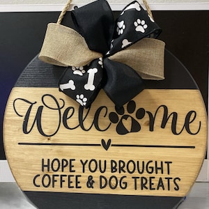 May include: Round wooden welcome sign with a black and natural wood finish. The sign features the word "Welcome" in a script font, paw prints, and the phrase "HOPE YOU BROUGHT COFFEE & DOG TREATS." A bow with paw prints and bone patterns adorns the top.