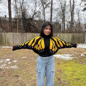 Puede incluir: Un bolero de ganchillo negro y amarillo con forma de alas de mariposa. El bolero presenta una base negra con diseños de alas amarillas y detalles de puntos blancos. La prenda se lleva sobre un cuello alto negro y se combina con vaqueros azul claro.