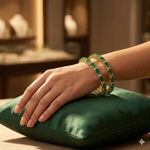 May include: Two gold bangles featuring a series of oval-cut green gemstones alternating with small clear stones. The bracelets are worn on a wrist resting on a dark green velvet cushion, showcasing a classic design with a polished metal finish.