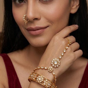 May include: A gold-toned hand harness featuring a wide, floral-patterned cuff bracelet connected by a delicate chain to a central flower motif and a ring. A matching gold-toned nose ring with small, clear, inset stones is also visible.