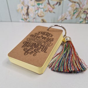 May include: A small brown notebook with a yellow edge and a colourful tassel keychain. The notebook has the text "There's always something to be thankful for" printed on the cover.