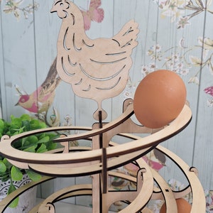 May include: Wooden egg holder shaped like a spiral with a chicken cutout on top. The holder is made of light brown wood and has a natural finish. There are several eggs in the holder, and one egg is rolling down the spiral.