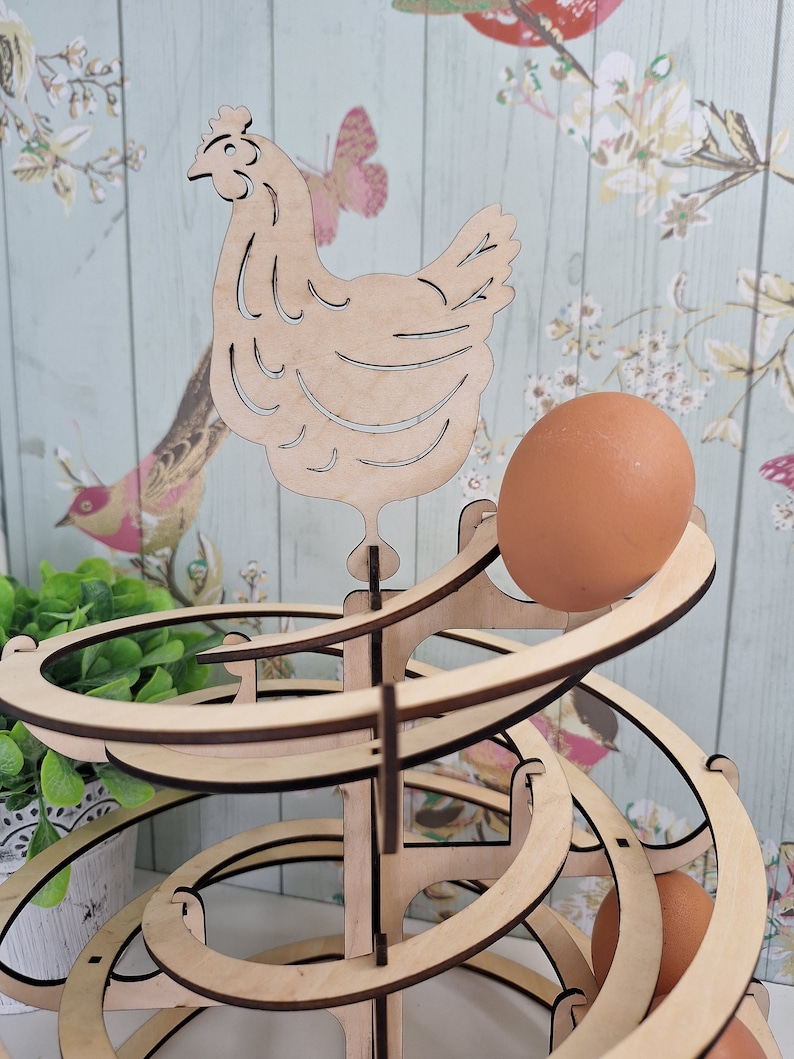 May include: Wooden egg holder shaped like a spiral with a chicken cutout on top. The holder is made of light brown wood and has a natural finish. There are several eggs in the holder, and one egg is rolling down the spiral.