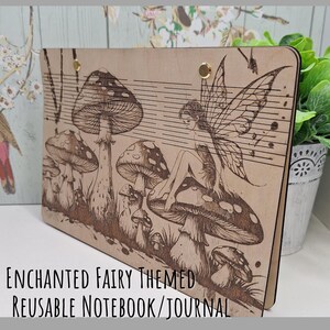 May include: A wooden notebook with an engraved fairy and mushroom design. The notebook has a light brown colour and features the text "ENCHANTED FAIRY THEMED REUSABLE NOTEBOOK/JOURNAL". The design includes musical notes and gold-coloured fasteners.