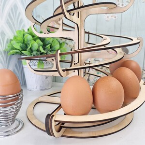 May include: Wooden egg holder shaped like a tree with a spiral design. The holder has four brown eggs on the bottom level and a single egg in a metal egg cup on a white surface.