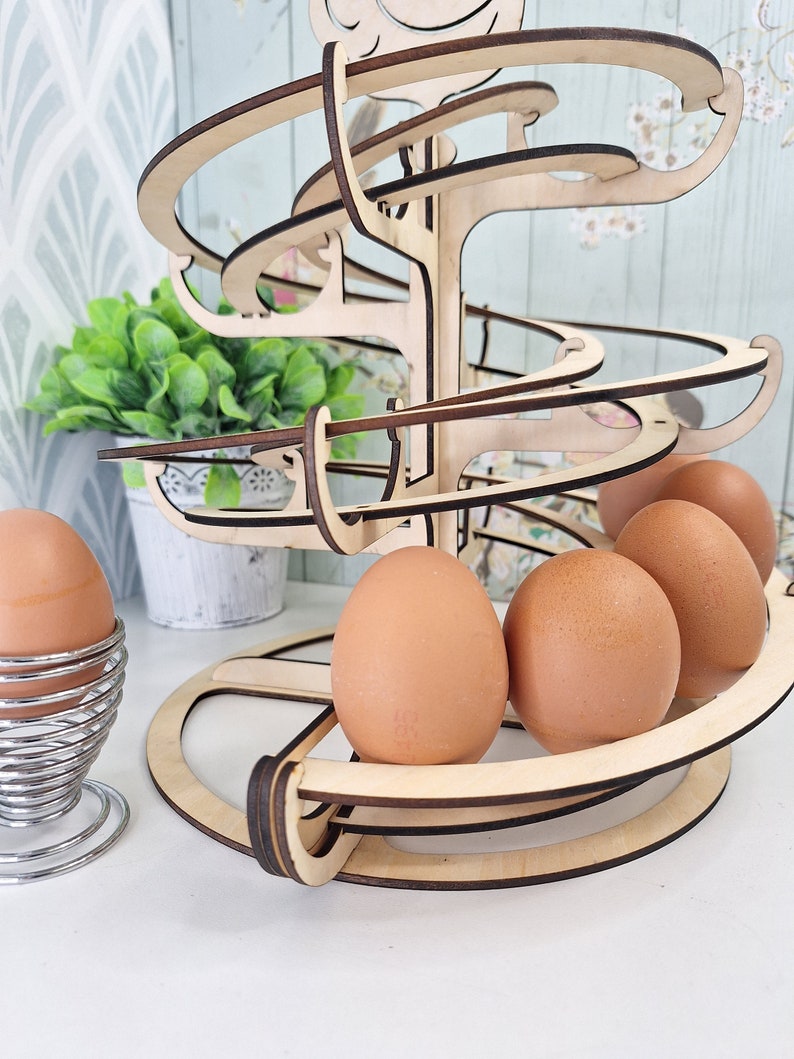 May include: Wooden egg holder shaped like a tree with a spiral design. The holder has four brown eggs on the bottom level and a single egg in a metal egg cup on a white surface.