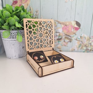 May include: A wooden box with a geometric pattern on the lid. The box is open and contains two pairs of earrings. One pair is iridescent and the other is white with a flower design.