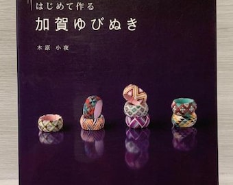 Puede incluir: Un libro titulado "Cómo hacer Kusa-shime" con una descripción de texto japonés. La portada presenta un fondo morado con cuatro dedal de costura coloridos, tres apilados y uno solo, sobre una superficie reflectante.