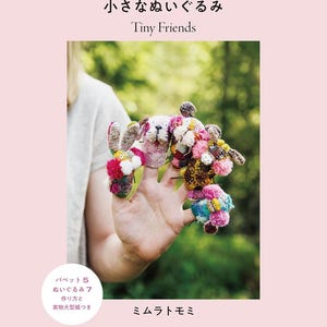 Può includere: Copertina di un libro con il titolo "Tiny Friends" in inglese e giapponese. L'immagine presenta diversi piccoli e colorati animali di peluche fatti a mano, tenuti nella mano di una persona. Lo sfondo è rosa tenue.