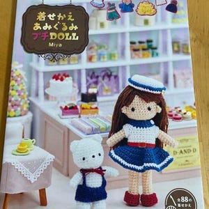 Könnte beinhalten: Buchcover mit einer Häkelpuppe und einem Bären, vor dem Hintergrund einer Bäckerei. Die Puppe trägt ein blau-weißes Matrosen-Outfit. Der Buchtitel lautet "着せかえ あみぐるみ プチ DOLL Miya".