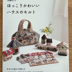 Puede incluir: Un libro titulado "House of Cozy Charm" con un título japonés encima. La portada del libro presenta una bolsa de patchwork, una variedad de pequeñas casas de patchwork y un estuche de lápices de patchwork. El texto de la portada del libro dice "House of Cozy Charm" y "35 proyectos de patchwork".