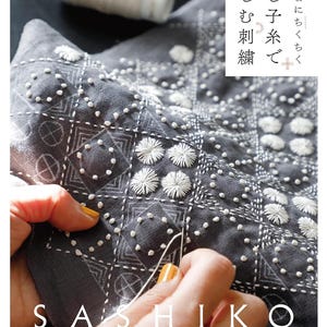 Könnte beinhalten: Nahaufnahme einer Hand, die einen grauen Stoff mit weißem Garn näht. Der Stoff hat ein geometrisches Muster und wird mit einer traditionellen japanischen Sticktechnik namens "Sashiko" gestickt. Der Text "SASHIKO" ist in Weiß auf den Stoff gedruckt.