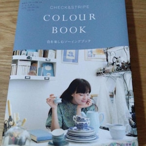Puede incluir: Un libro titulado "COLOUR BOOK" de Check & Stripe, con una cubierta azul claro. La imagen presenta a una mujer con cabello largo y oscuro, sentada en una mesa con porcelana azul y blanca. El título del libro está en mayúsculas blancas.