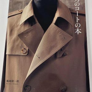 以下が含まれることがあります： 大きな襟と2つのボタンが付いた茶色のトレンチコート。画像の右側に「男のコートの本」という文字が印刷されています。