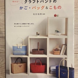 以下が含まれることがあります： 「クラフトバンドのかご・バッグ＆こもの」というタイトルの日本語の本の表紙。さまざまな色とスタイルの手作りのバスケットとバッグが棚に並べられています。