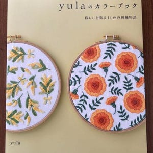May include: A book titled "yula の カラーブック" with the subtitle "暮らしを彩る 14 色の刺繍物語". The cover features two embroidery hoops with floral designs. One hoop has yellow flowers and green leaves, while the other has orange flowers and green leaves.