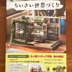 Może przedstawiać: Okładka książki z żółtym tłem i tytułem "ちいさい世界づくり" (Chiisai Sekai-zukuri) w dużych czarnych literach. Książka opowiada o tworzeniu miniaturowych światów, czyli dioram, z materiałów dostępnych w domu. Okładka przedstawia miniaturowy domek z zielonym dachem i małym ogrodem.